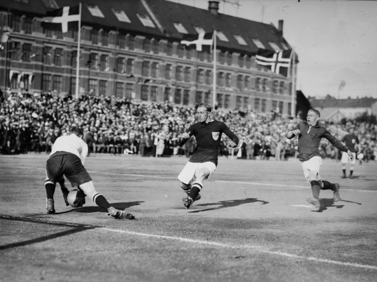 Fotball. Keeper griper ballen langs bakken. To angrepsspillere løper mot ham. Norske og danske flagg.