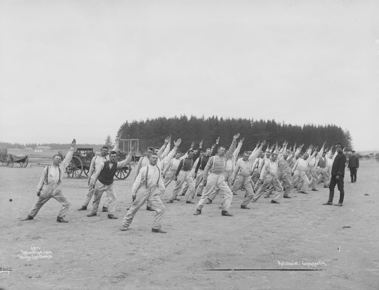 Str gruppe menn løfter venstre arm i været, bøy i venstre ben. Hest og kjerre i bakgrunnen, uniformert instruktør til høyre.
