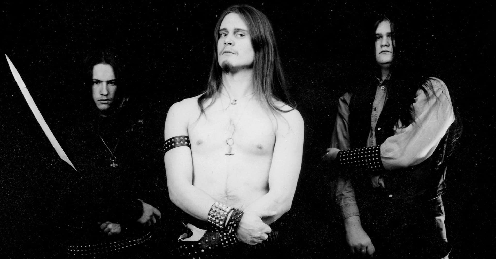 Halvtotal av tre skumle menn i heavy metal-klær ser skummelt inn i kamera. Den midterste står i bar overkropp.