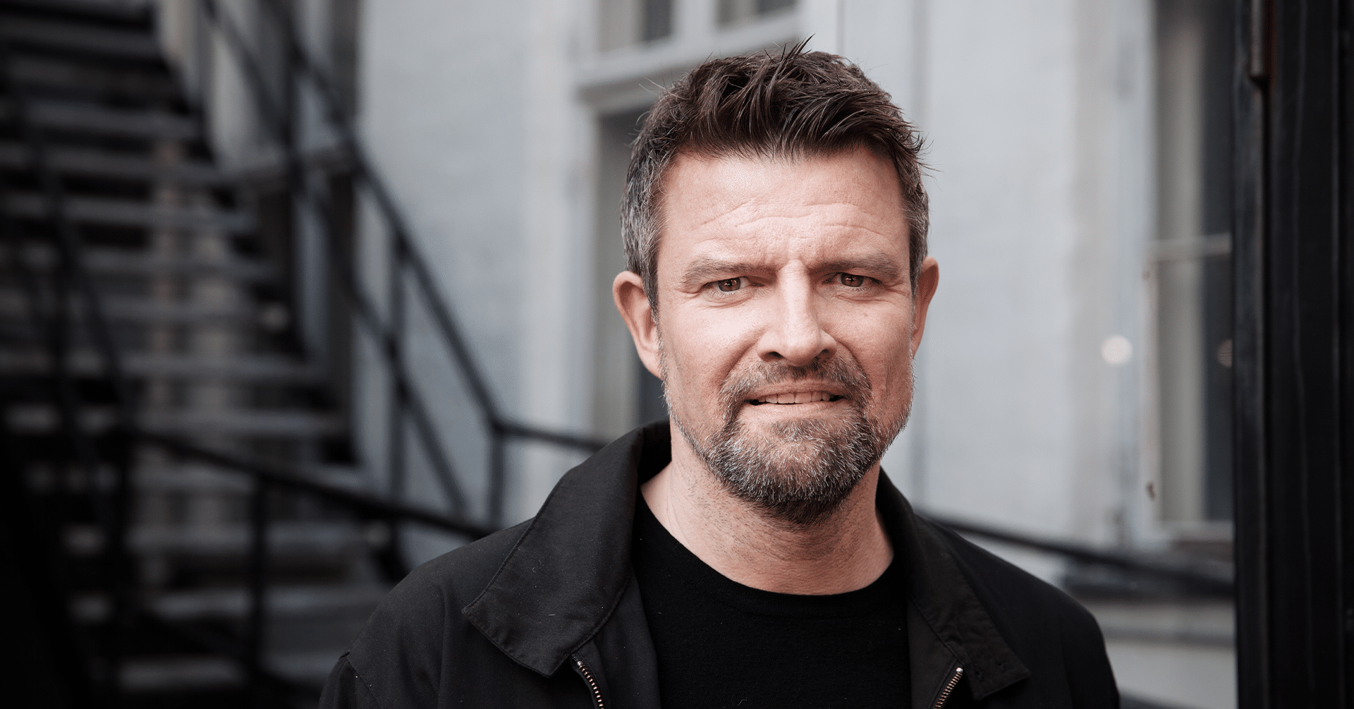 En mann med litt skjegg står foran en trapp og smiler mot kamera