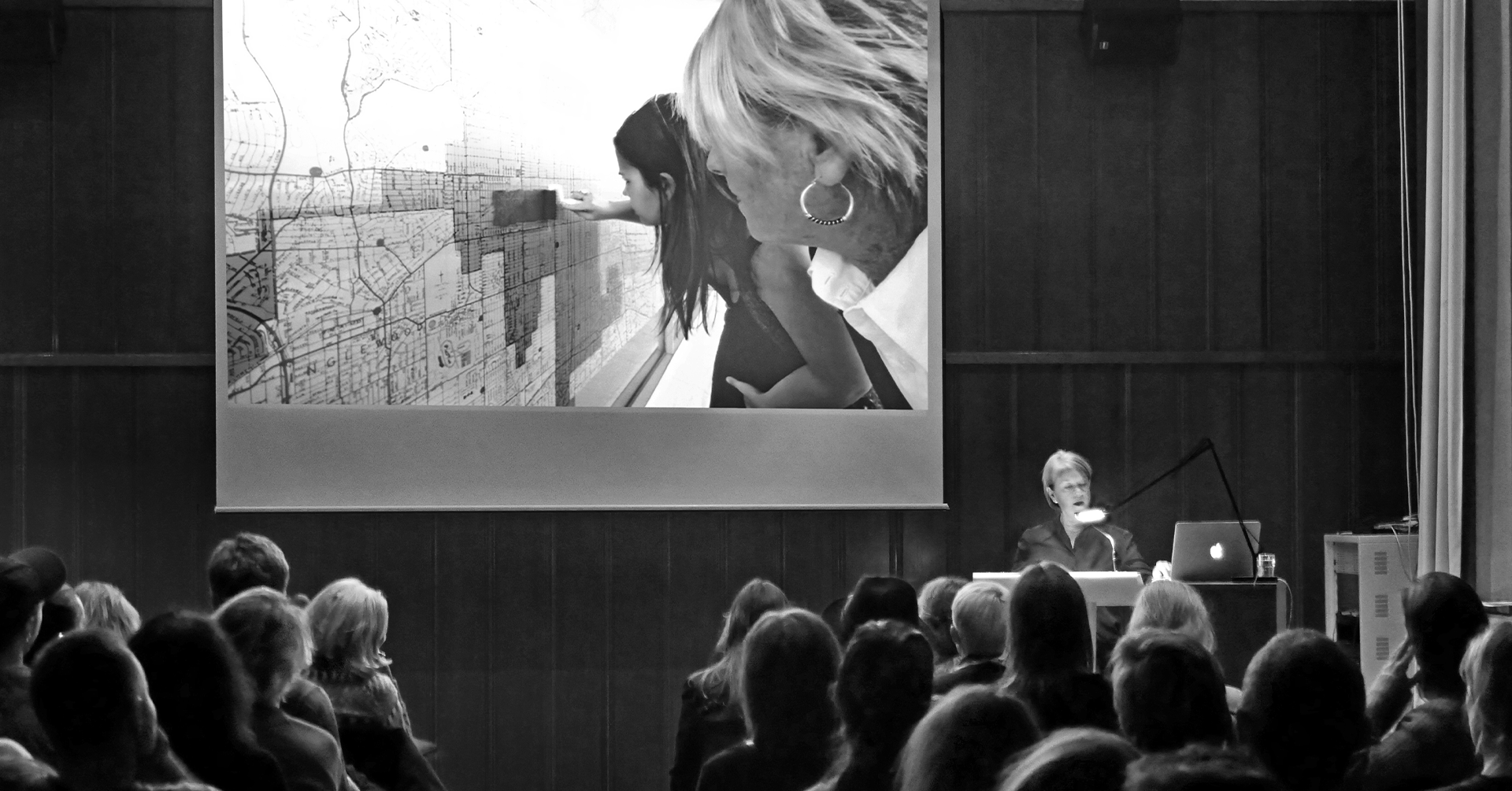 En sal med tilskuere ser på en fremvisning på et lerret og en kvinne snakker fra en talerstol.