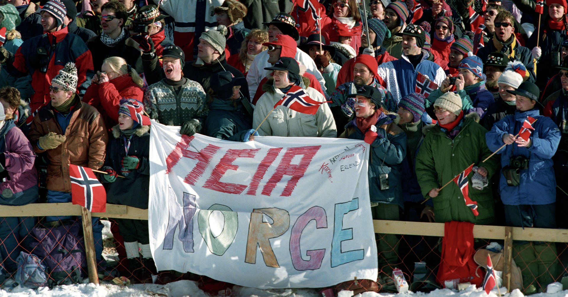 En stor gruppe mennesker samlet sammen for å heie. Det er vinter og noen holder et stort banner med "Heia Norge" på.