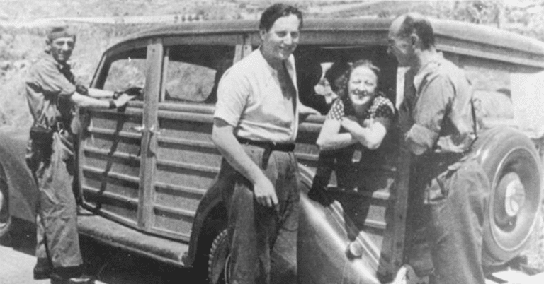 En kvinne sitter i en gammel bil og smiler mot kameraet. Tre menn står utenfor bilen, to smiler.
