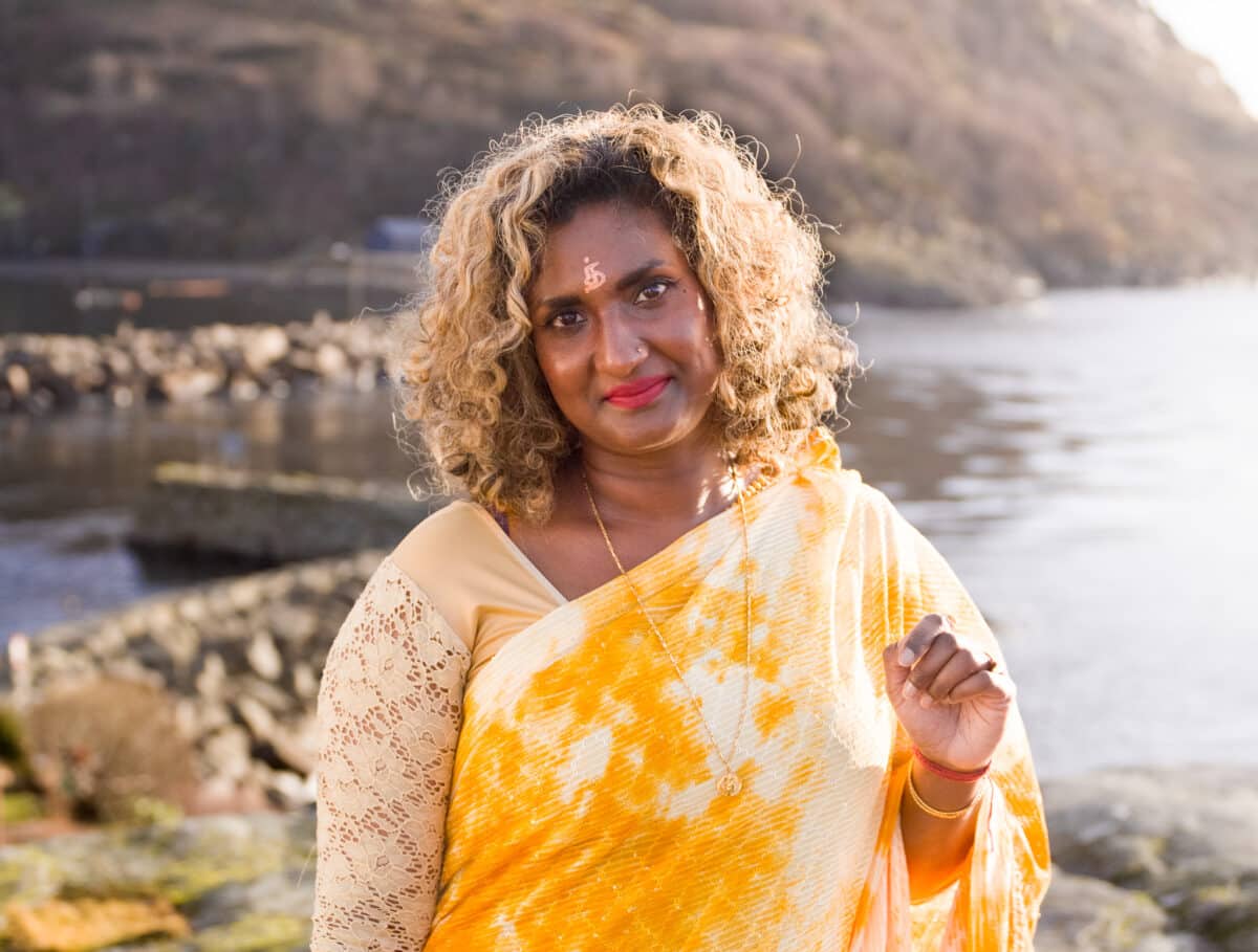 Melaninrik kvinne med gul sari stor foran et kystlandskap i sol. Smiler mot kameraet. Fotografi.