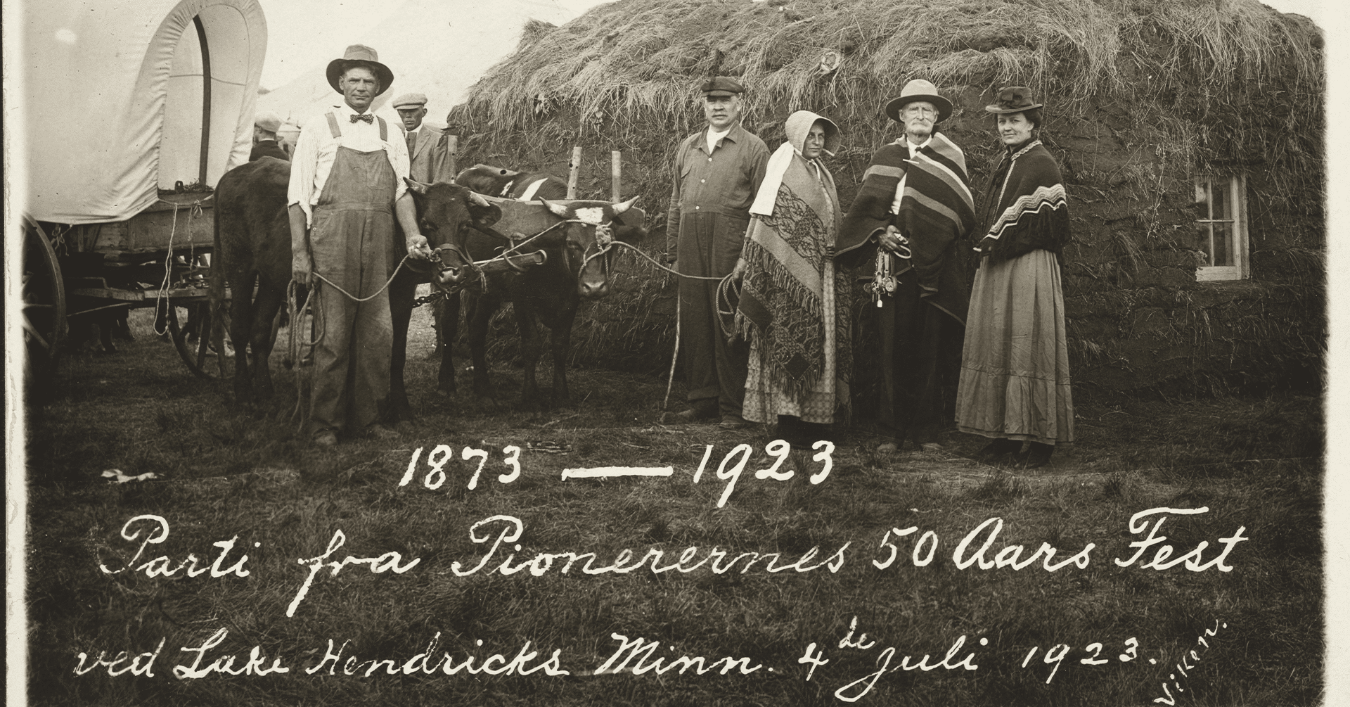 Et følge på kvinner og menn i ført overaller, hatt og poncho. Mennene holder i hver sin okse som drar en conestoga vogn. De poserer foran et hus som består av leire og strå.