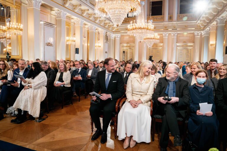 A formal event inside the Royal Palace featuring an opulent hall with tall white columns, gilded details, and multiple large crystal chandeliers hanging from the ceiling. The room is filled with rows of seated attendees dressed in formal attire. In the foreground, four individuals are seated in ornate wooden chairs: the Norwegian Crown Prince and Crown Princess, author Jon Fosse, and his wife, Anna Fosse.