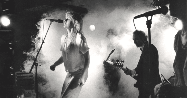 En kvinne synger fra en scene, hun har på jeans og hvit t-skjorte. Det er mye røyk på scenen og man kan se gitaristen i bakgrunnen.
