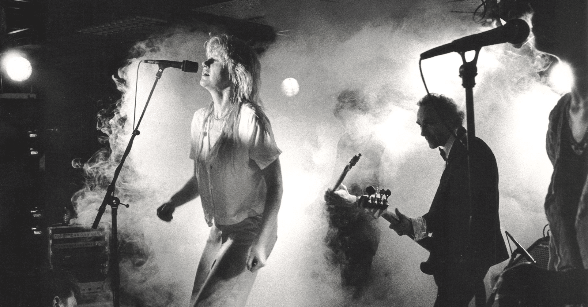 En kvinne synger fra en scene, hun har på jeans og hvit t-skjorte. Det er mye røyk på scenen og man kan se gitaristen i bakgrunnen.