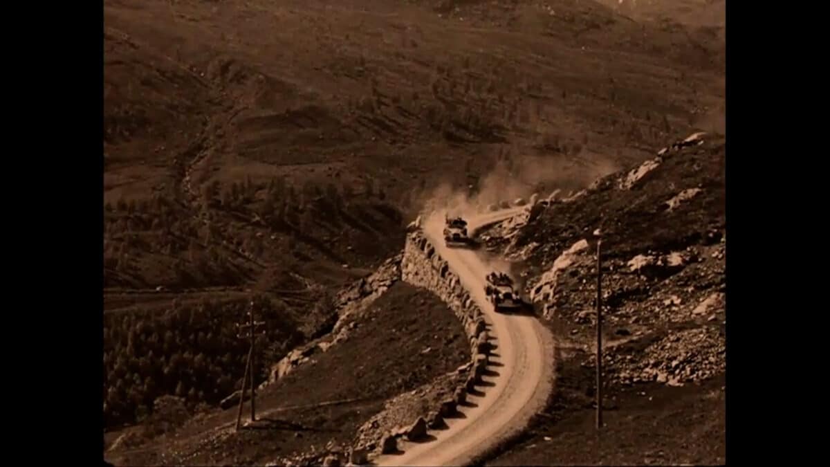 Gammelt svart-hvitt bilde i en brunlig tone av to biler som kjører på en gruset fjellvei