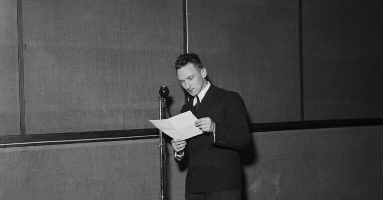 En mann i dress leser fra et ark foran en mikrofon på stativ