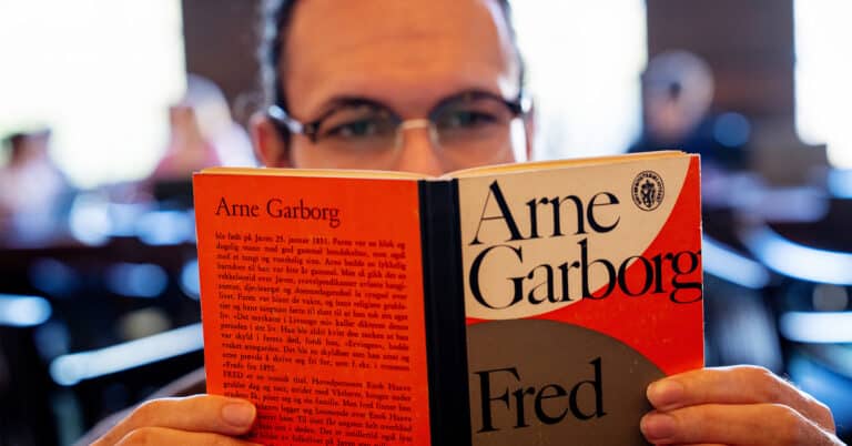 En nært fotografi av en person som holder en bok foran ansiktet sitt. Bokens omslag er rødt og grått, og den har tittelen 'Fred' skrevet i store svarte bokstaver sammen med forfatterens navn, Arne Garborg.