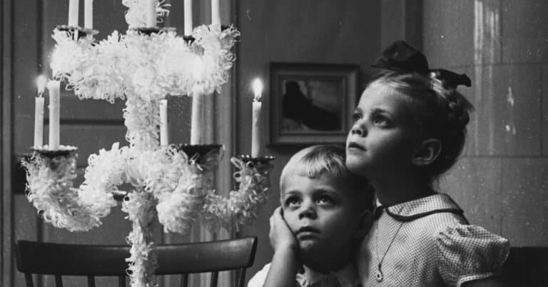 To små barn ser opp på en lysestake som er pyntet til jul. Barna er veldig pent kledd.