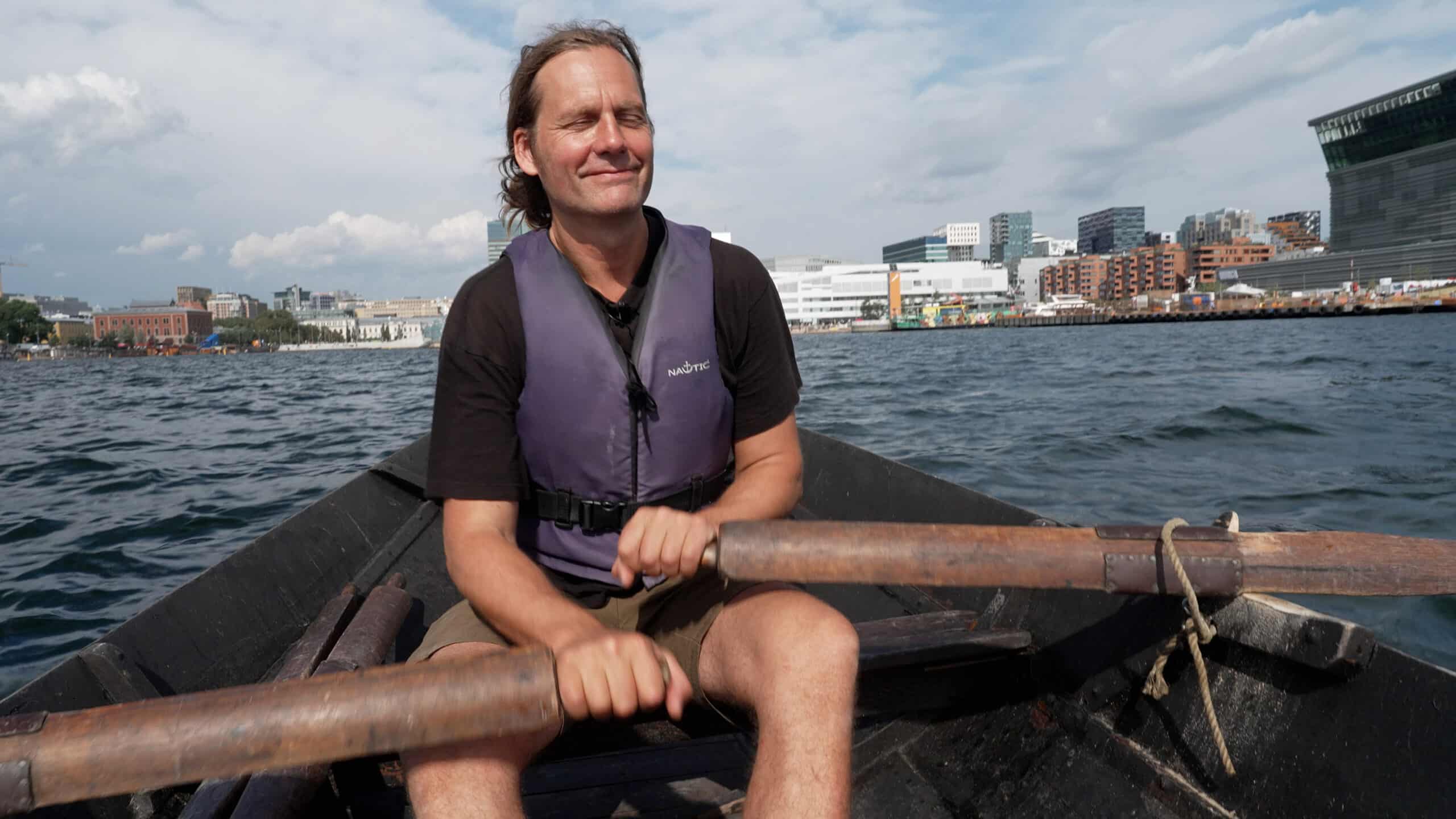 Person med lilla redningsvest merket «NAUTIC» ror en båt med treårer på vannet. Ansiktet er uskarpt, og i bakgrunnen sees både moderne og eldre bygninger langs vannkanten under delvis skyet himmel.