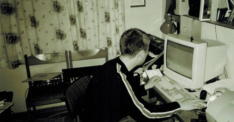 Person sitter ved et skrivebord og bruker en stasjonær datamaskin. Bildet er i sort-hvitt og viser et typisk hjemmemiljø fra 1990-tallet, med gardiner med mønster, en bordlampe og papirer på pulten. Personen ser mot skjermen og har hendene på tastaturet.