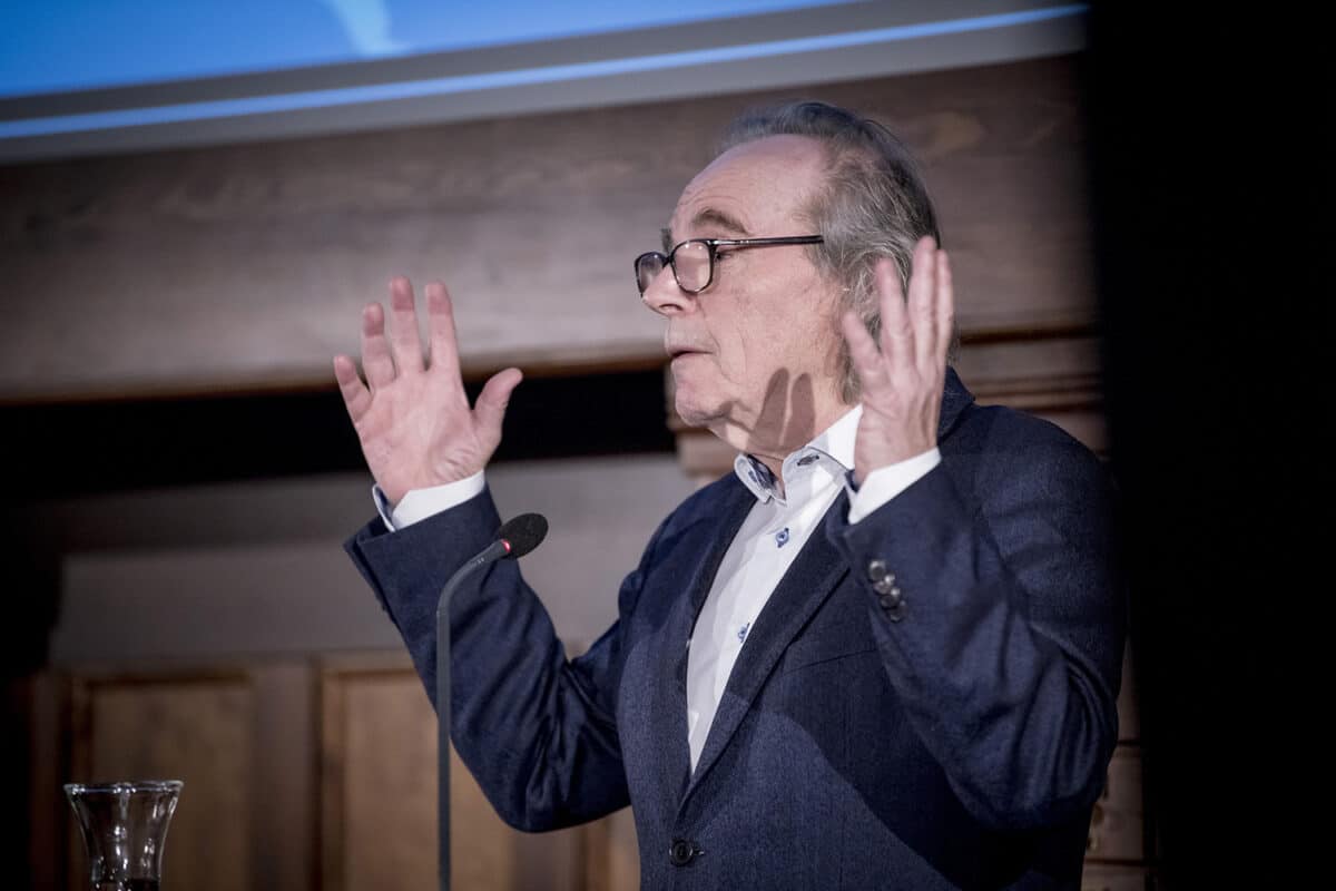 En person med grått hår, briller og mørk dressjakke står ved en talerstol og holder et foredrag. Personen gestikulerer med hendene løftet. I forgrunnen står et mikrofonstativ.