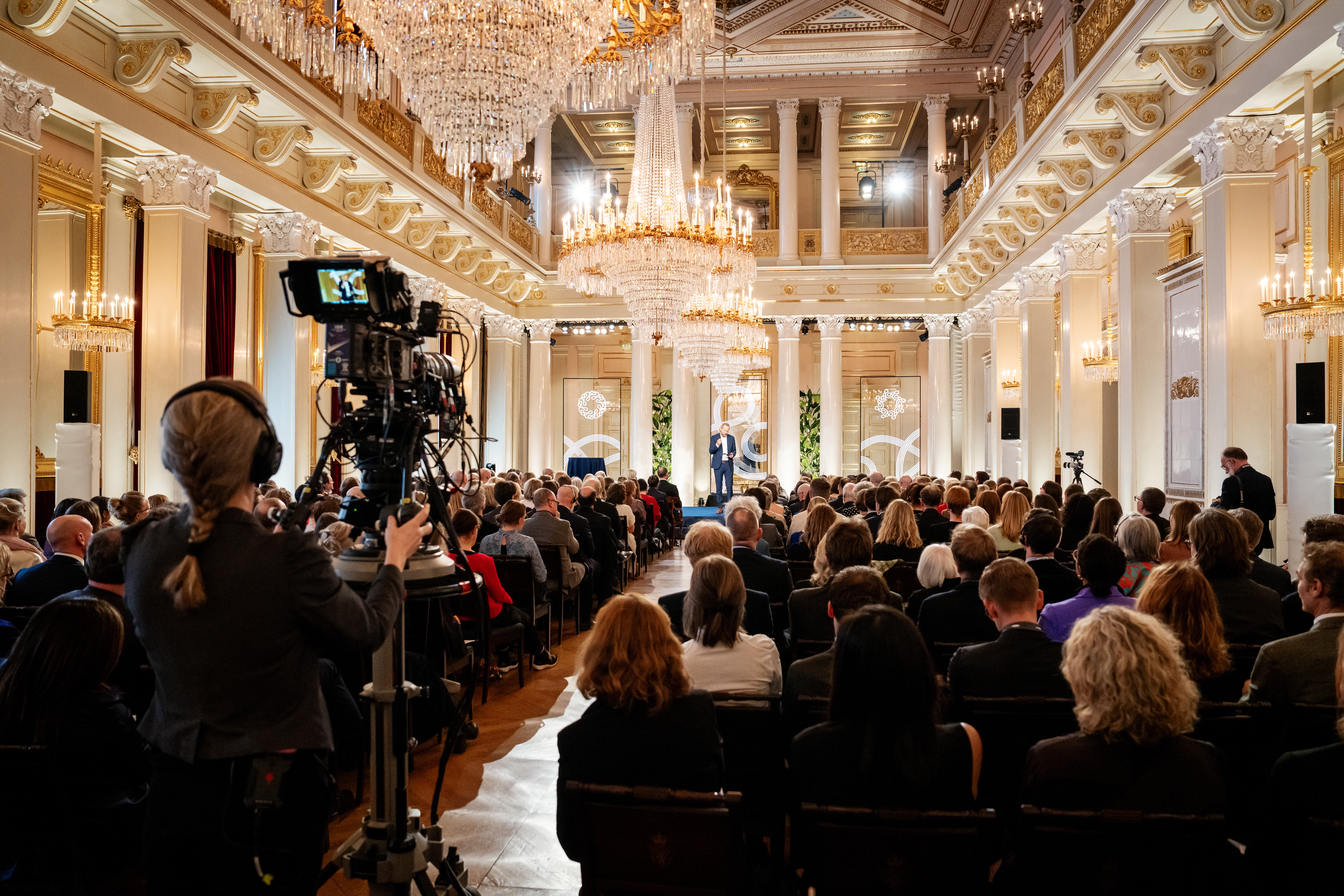 A large audience seated in rows inside the Royal Palace, facing a stage where a speaker stands. A camera operator is filming the event from the side. The setting is formal, with bright lighting and chandeliers visible above.