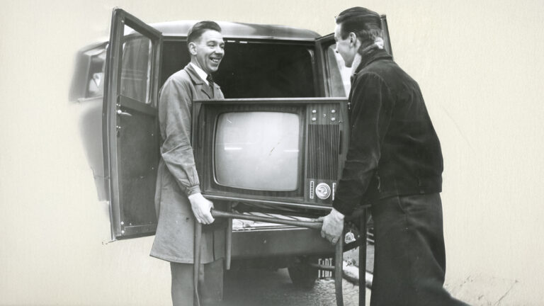 To personer løfter en stor, gammeldags fjernsynsapparat med trebenet stativ inn eller ut av en bil med åpne bakdører. TV-en har avrundet skjerm, dreieknapper og ventilasjonsriller på siden, typisk for 1960-tallets design.