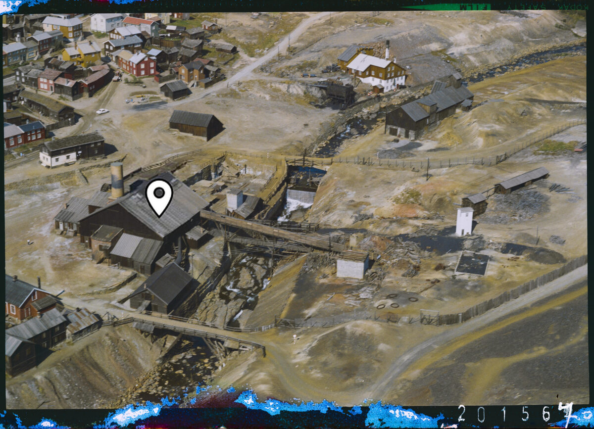 Flyfoto av et historisk gruveområde med trebygninger og stier i et nakent landskap. I forgrunnen ligger en stor bygning med pipe og flere trebroer som forbinder bygningene. Rundt området sees mindre hus, gjerder og en bekk som slynger seg gjennom terrenget. Et hvitt tårn står til høyre i bildet. Bildet har et markert punkt på taket til den største bygningen.