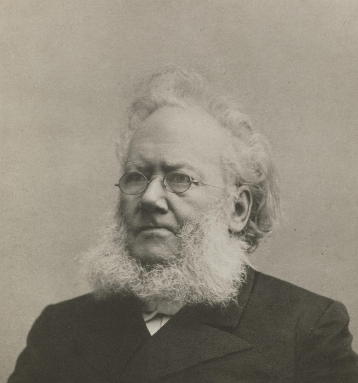 Fotografi av Henrik Ibsen. Hvit mann med hvitt hår og skjegg. Runde, små briller. Frakk. 