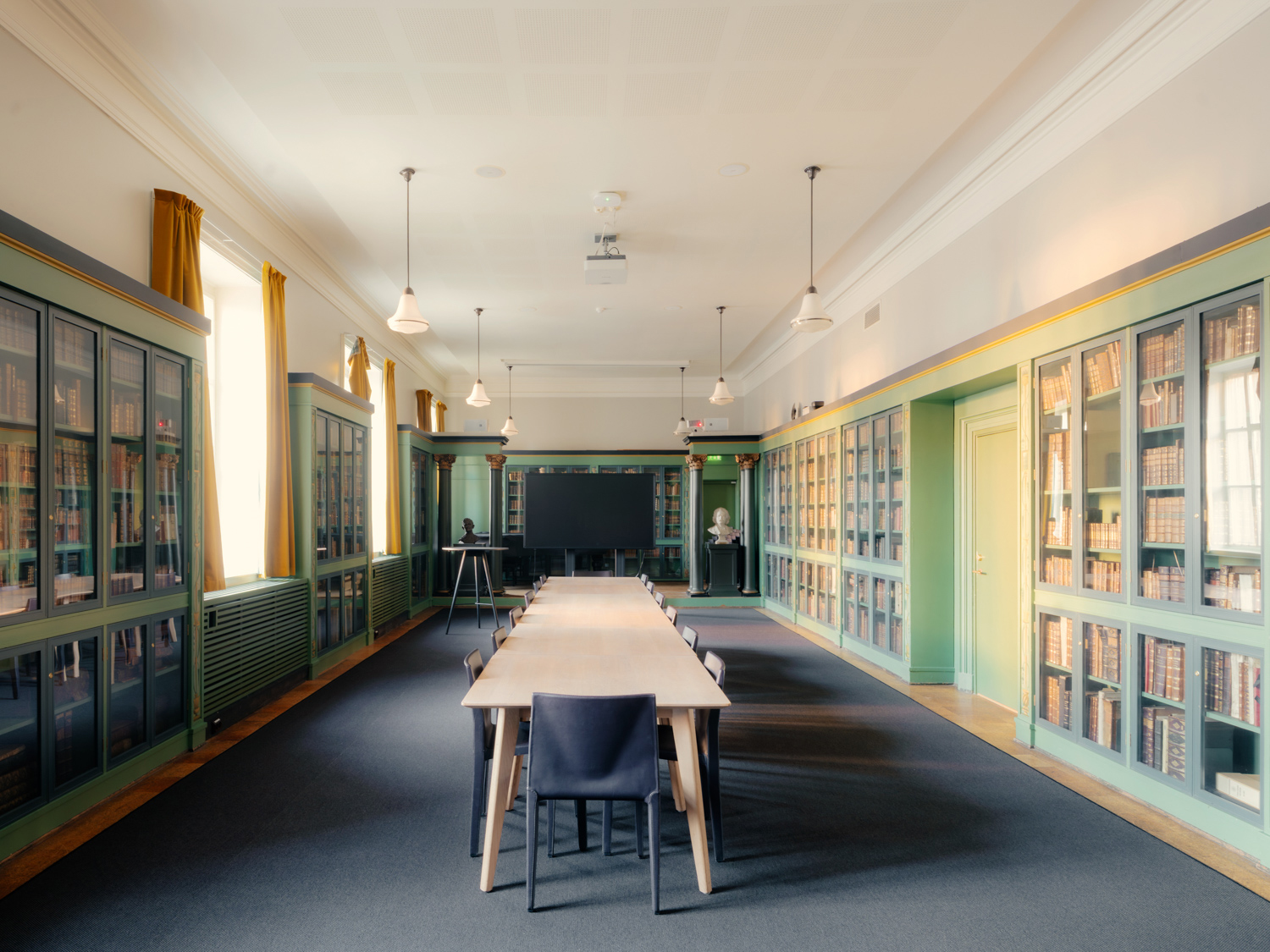Et langt møtebord omgitt av stoler i et bibliotekrom med grønne bokhyller langs veggene og en stor skjerm i enden av rommet.