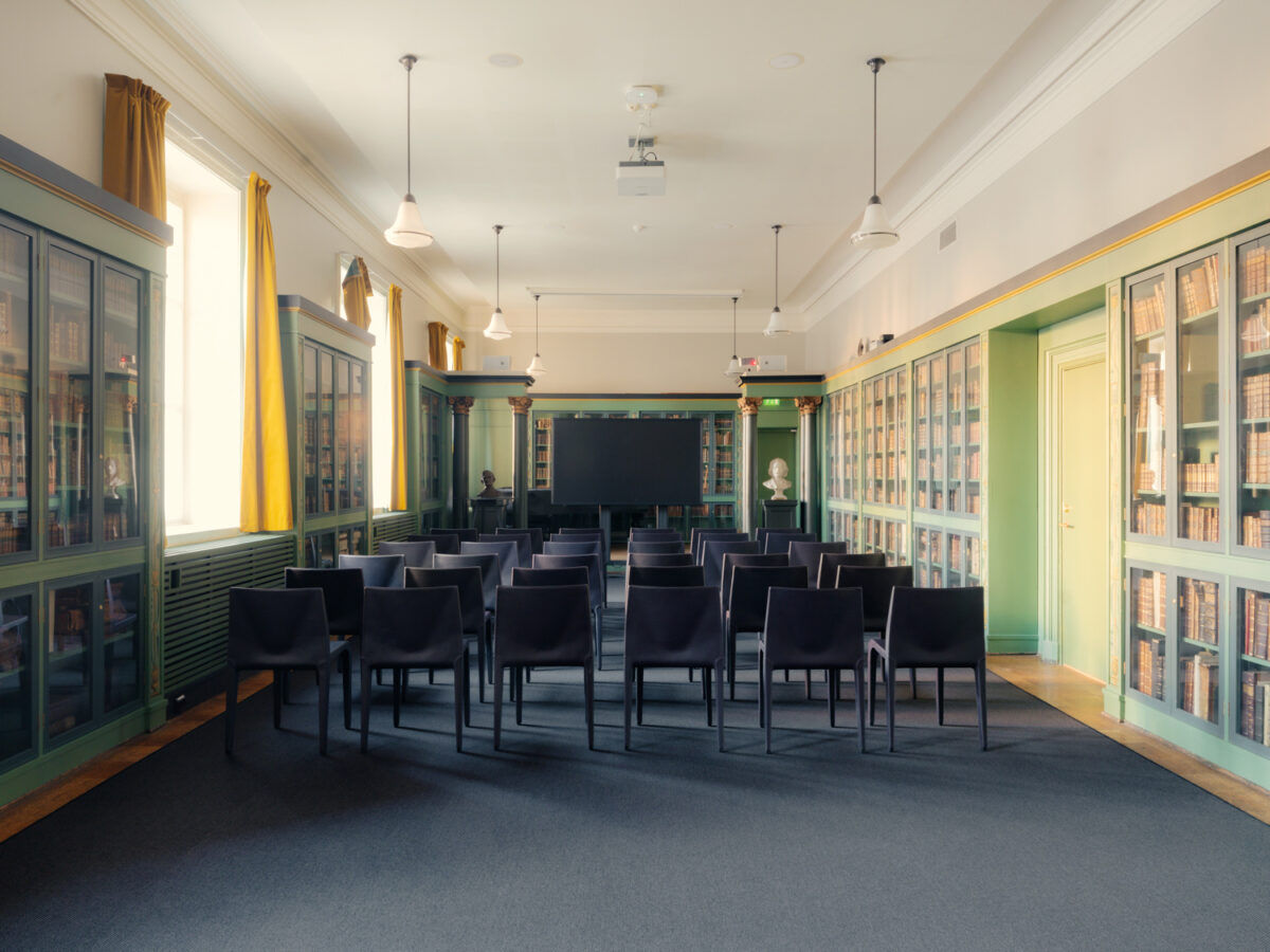 Et bibliotekrom med svarte stoler satt opp i et auditorium‑oppsett, omgitt av grønne bokhyller og store vinduer med gule gardiner.