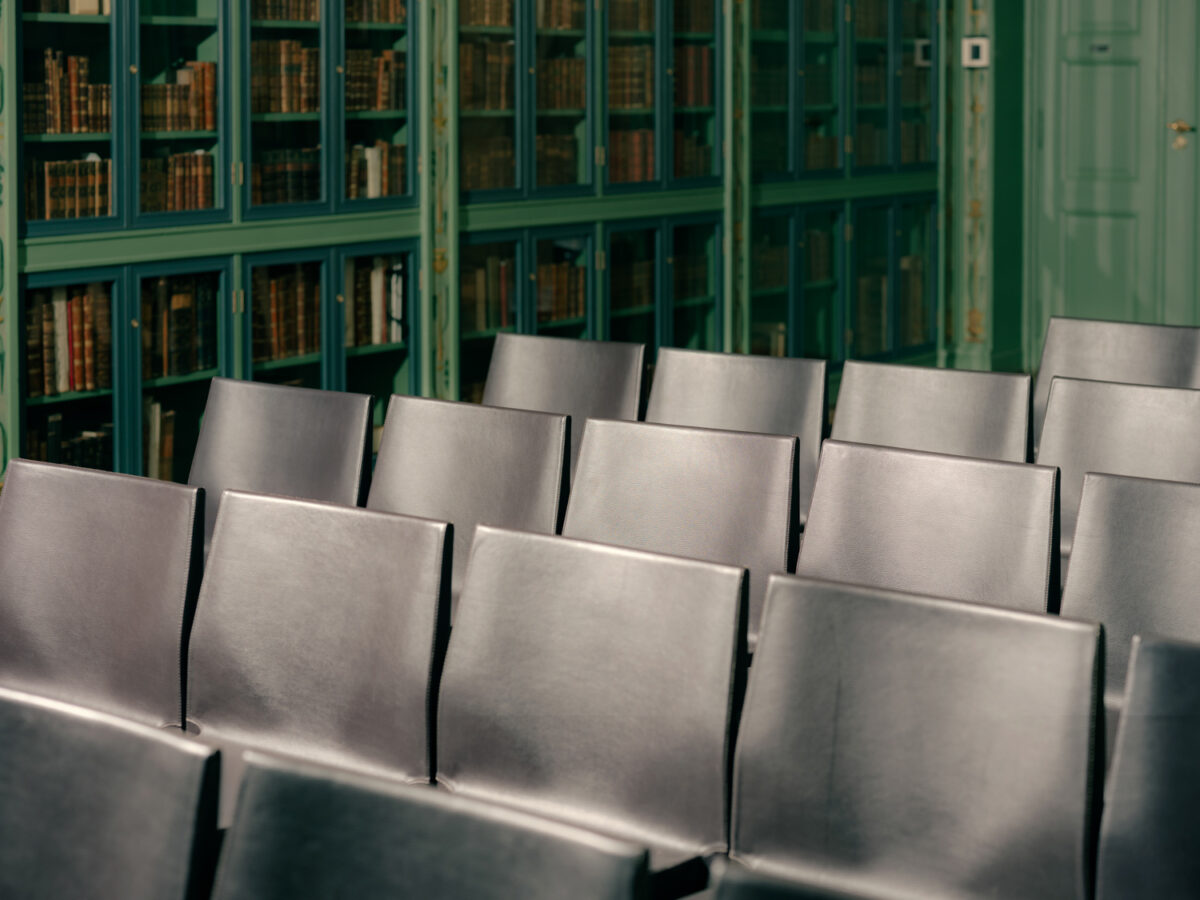 Rader med svarte stoler foran grønne bokhyller fylt med gamle bøker i et klassisk bibliotekrom.