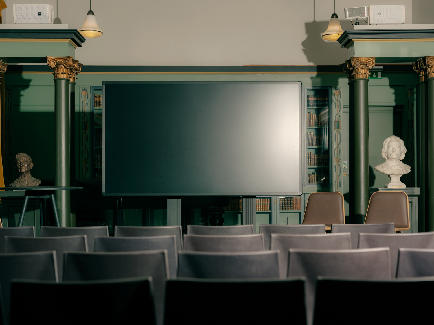 En stor skjerm plassert foran rader med stoler i et klassisk bibliotekrom, flankert av byster på sokler og grønne søyler.