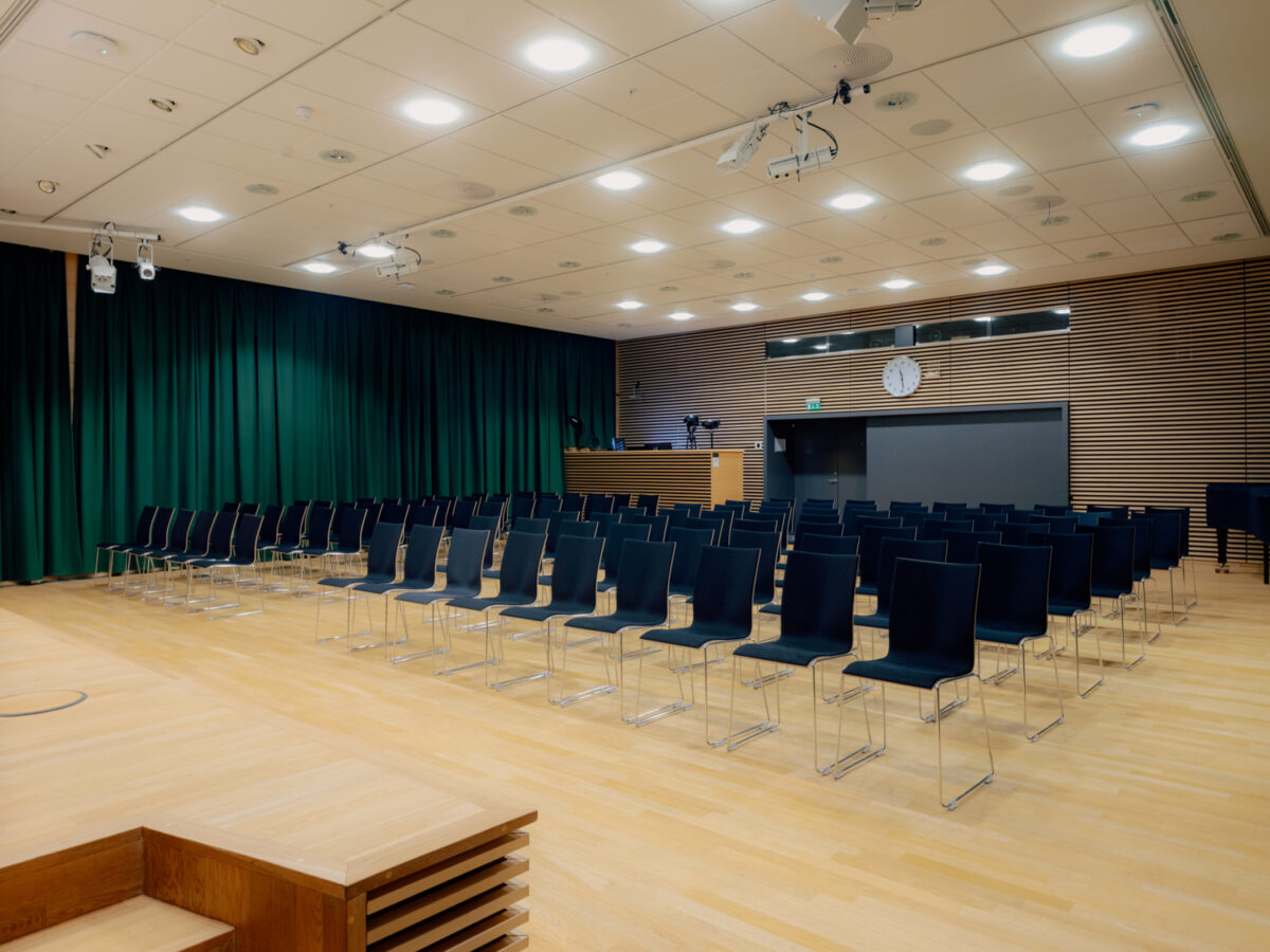 Et stort auditorium med lyse tregulv, svarte stoler i rekker og en liten scene foran grønne scenetepper.