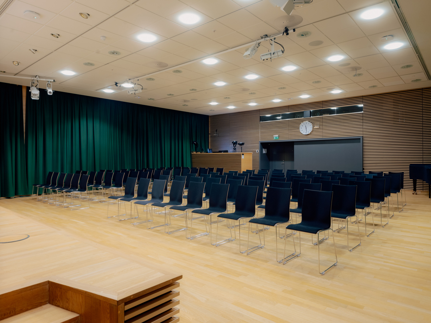 Et stort auditorium med lyse tregulv, svarte stoler i rekker og en liten scene foran grønne scenetepper.