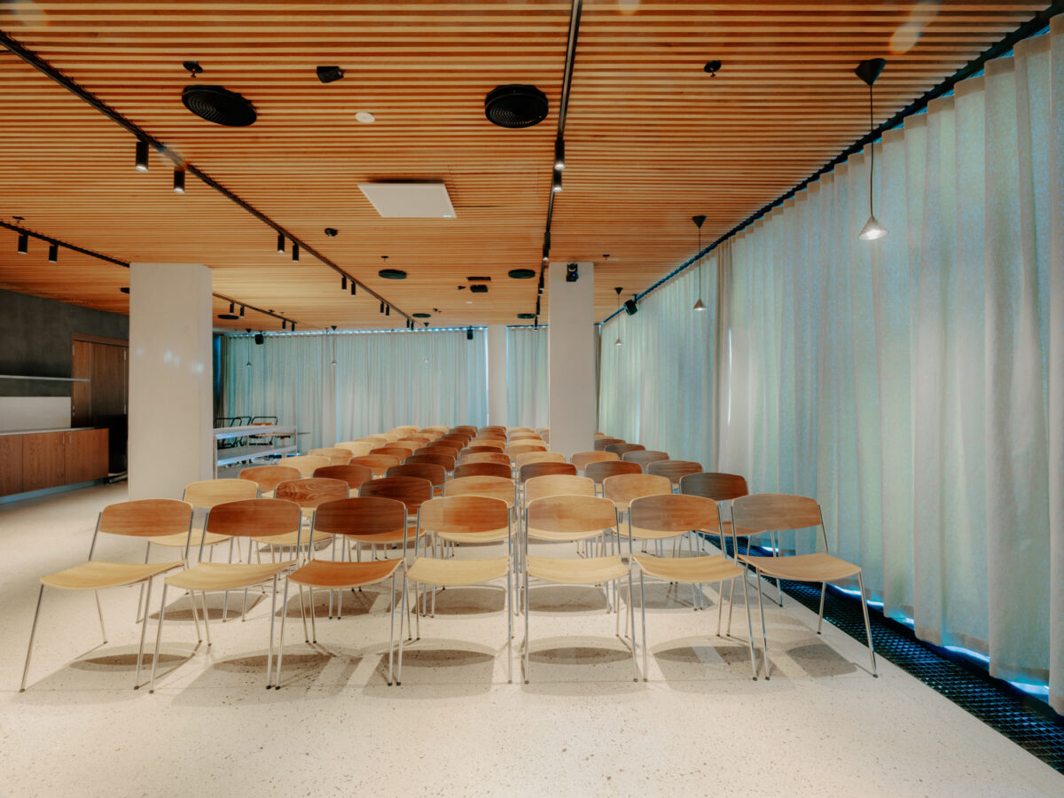 Konferanserom med flere rekker av trestoler og et høyt bord med to stoler i høyre side, omgitt av lys gjennom store gardiner.