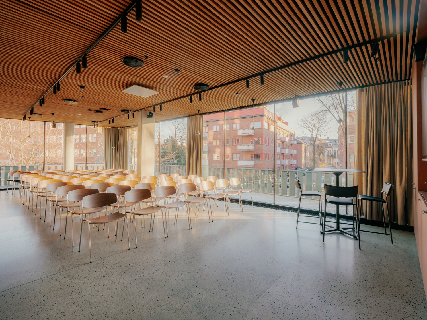Et lyst konferanserom med store vindusflater og rekker av trestoler satt opp i auditorium‑oppsett, med utsikt mot boligblokker utenfor.