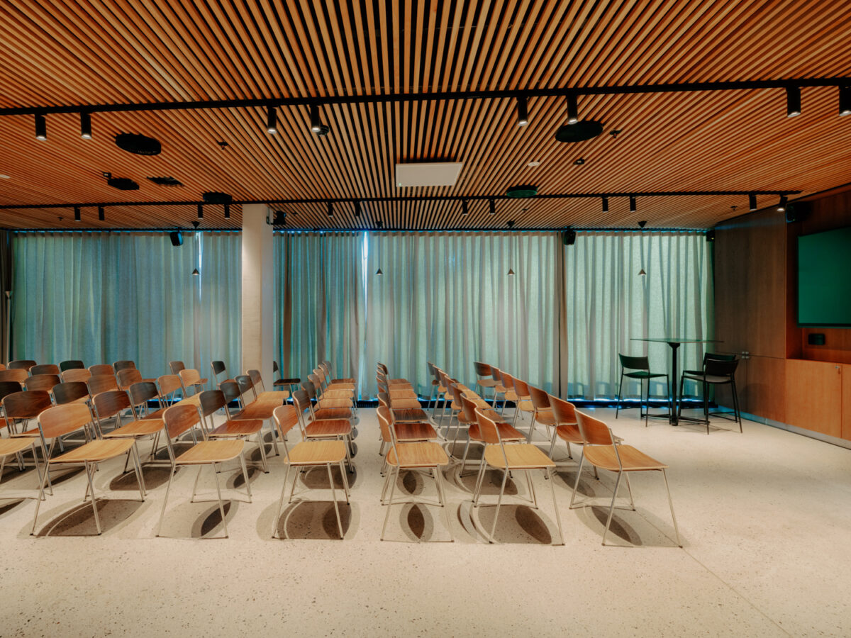 Konferanserom med flere rekker av trestoler vendt mot en skjerm og et høyt bord med to stoler langs siden av rommet.