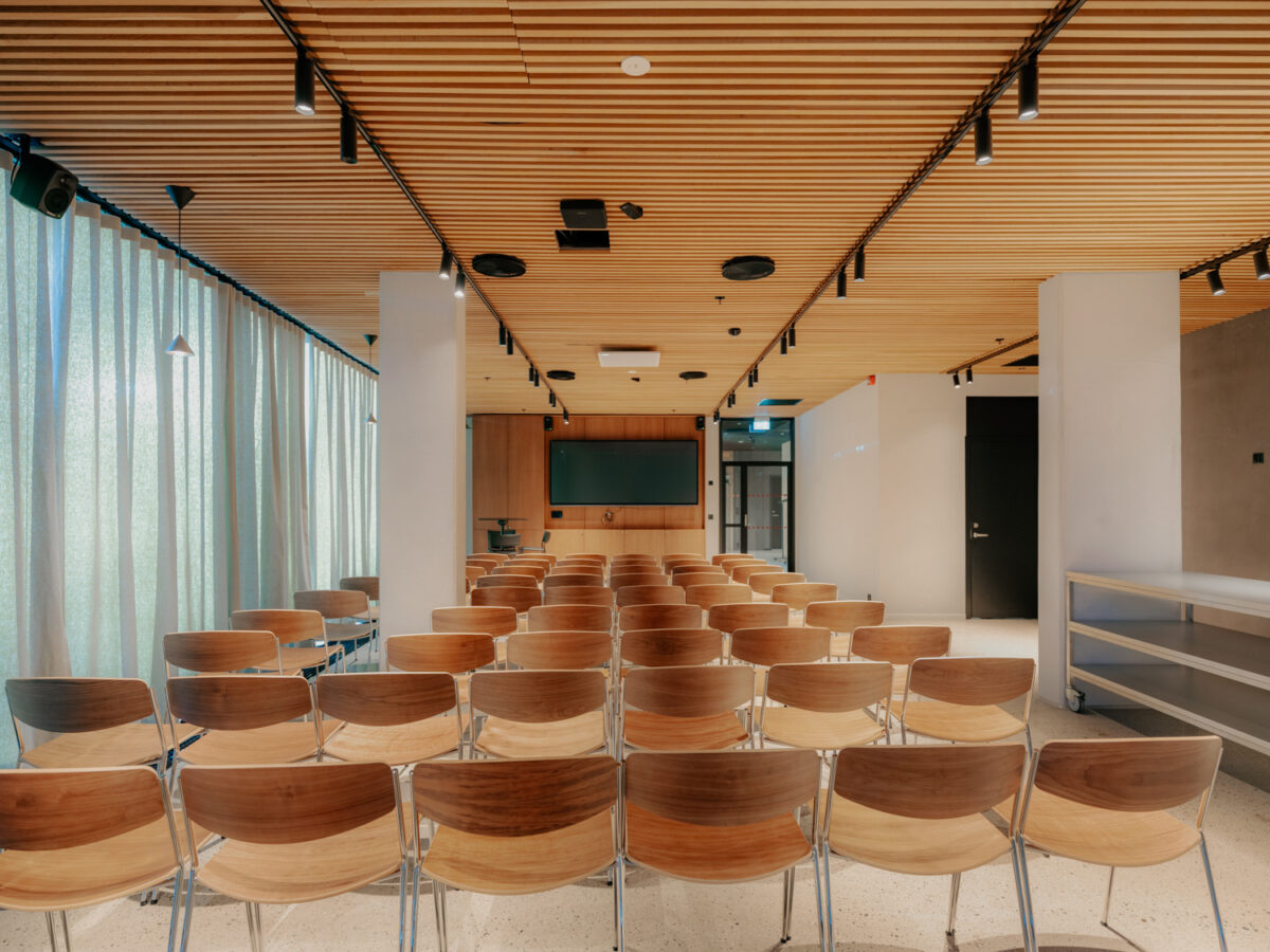 Rekker av trestoler plassert i et konferanserom med trepanel i taket, gardiner langs veggene og en skjerm i front.