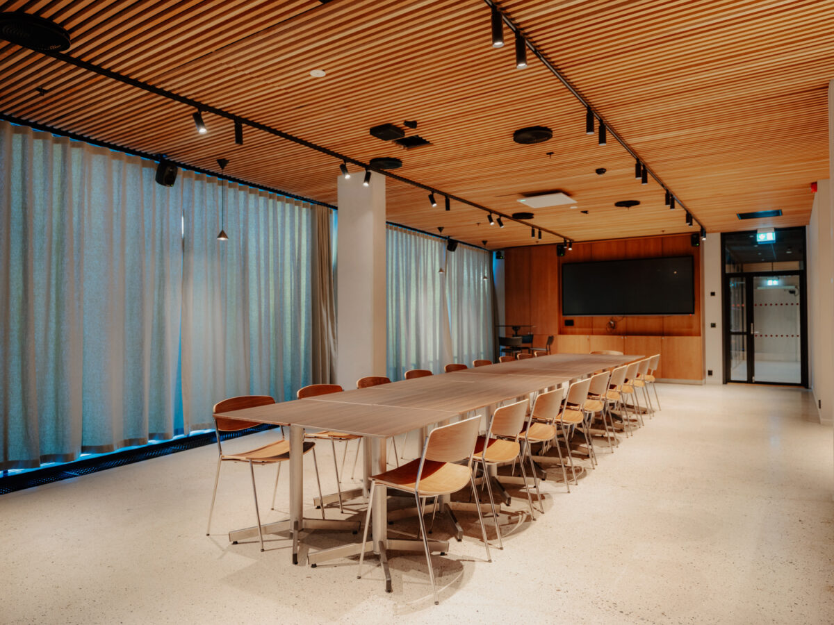 Et moderne møterom med en lang konferansebordrekke omgitt av trestoler, gardiner langs veggene og en stor skjerm i enden av rommet.