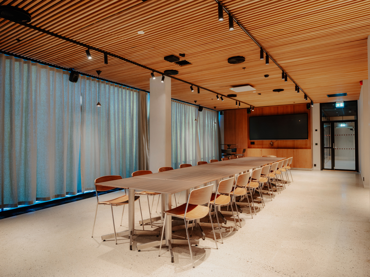 Et moderne møterom med en lang konferansebordrekke omgitt av trestoler, gardiner langs veggene og en stor skjerm i enden av rommet.