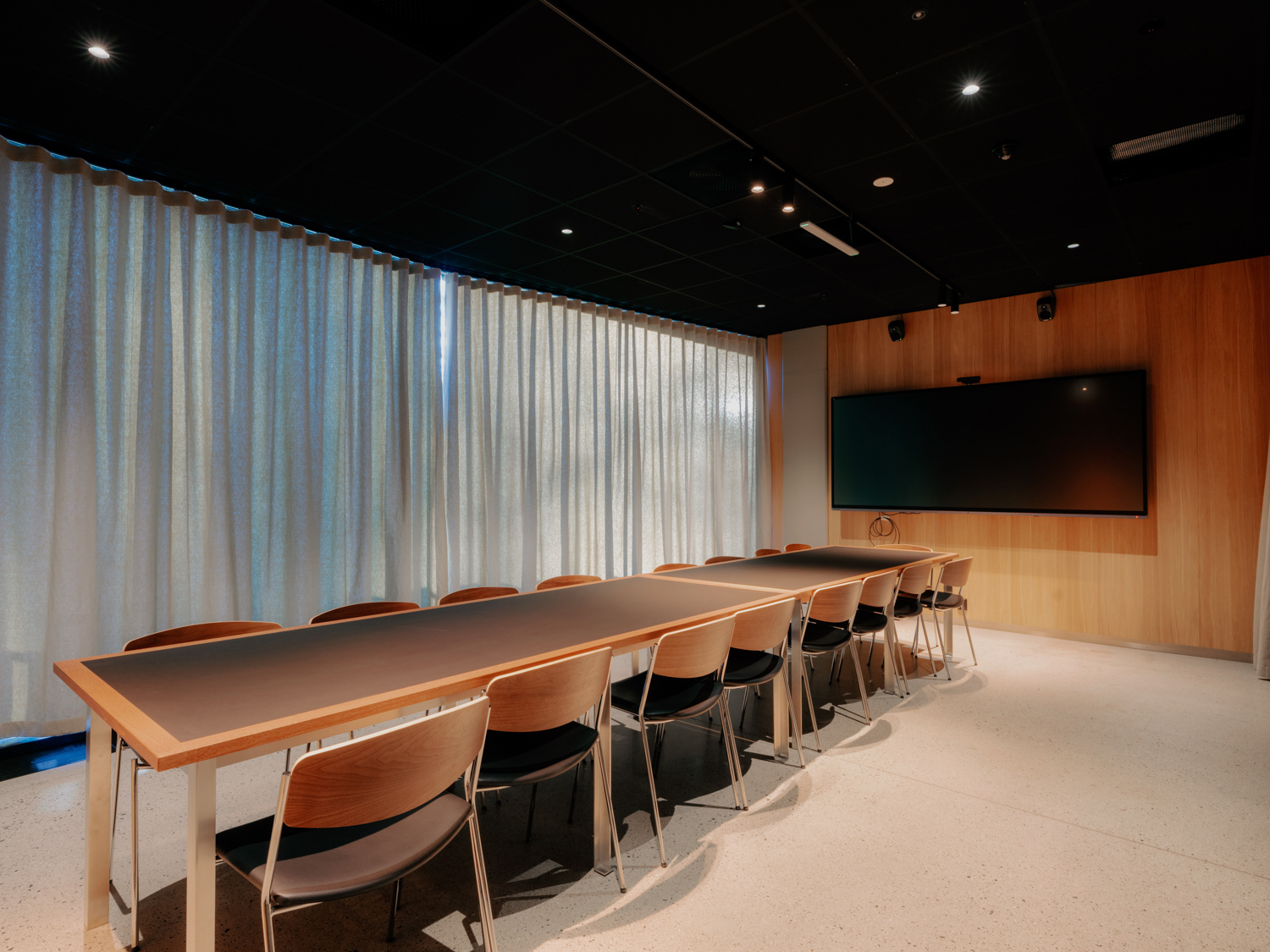 Et moderne møterom med et langt konferansebord omgitt av trestoler, lukkede gardiner langs veggene og en stor skjerm montert på en trevegg.
