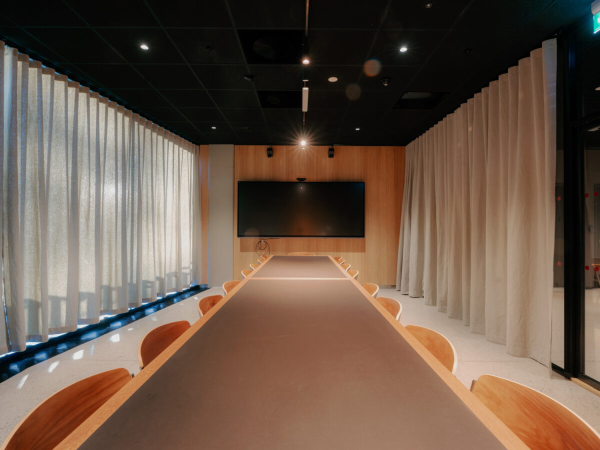 Et møterom sett fra enden av et langt konferansebord, med gardiner rundt rommet og en stor skjerm på veggen foran.