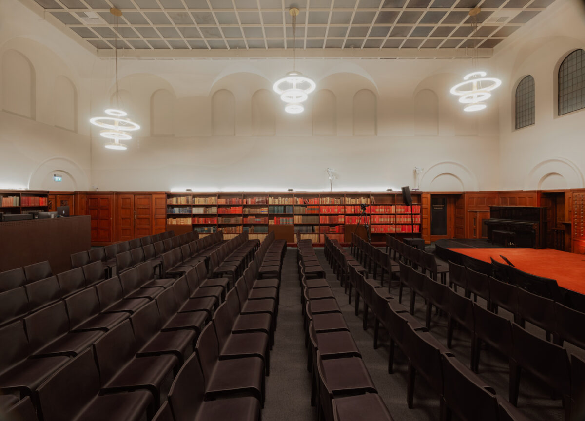 Et auditorium med mørke stoler i rekker, store bokhyller langs veggene og moderne runde lysekroner hengende fra taket.