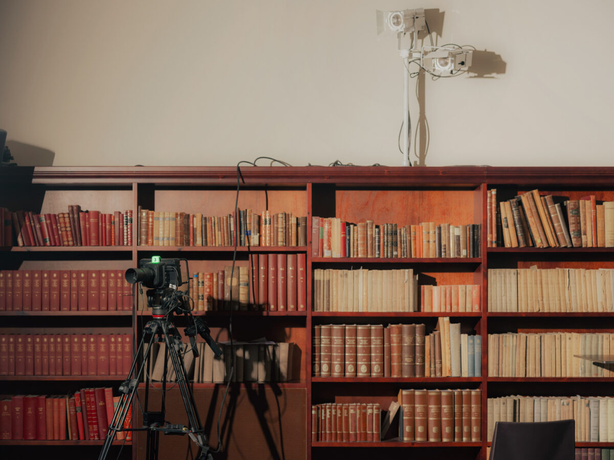 Et kamera på stativ står foran en stor bokhylle fylt med gamle bøker i et bibliotekslignende rom.