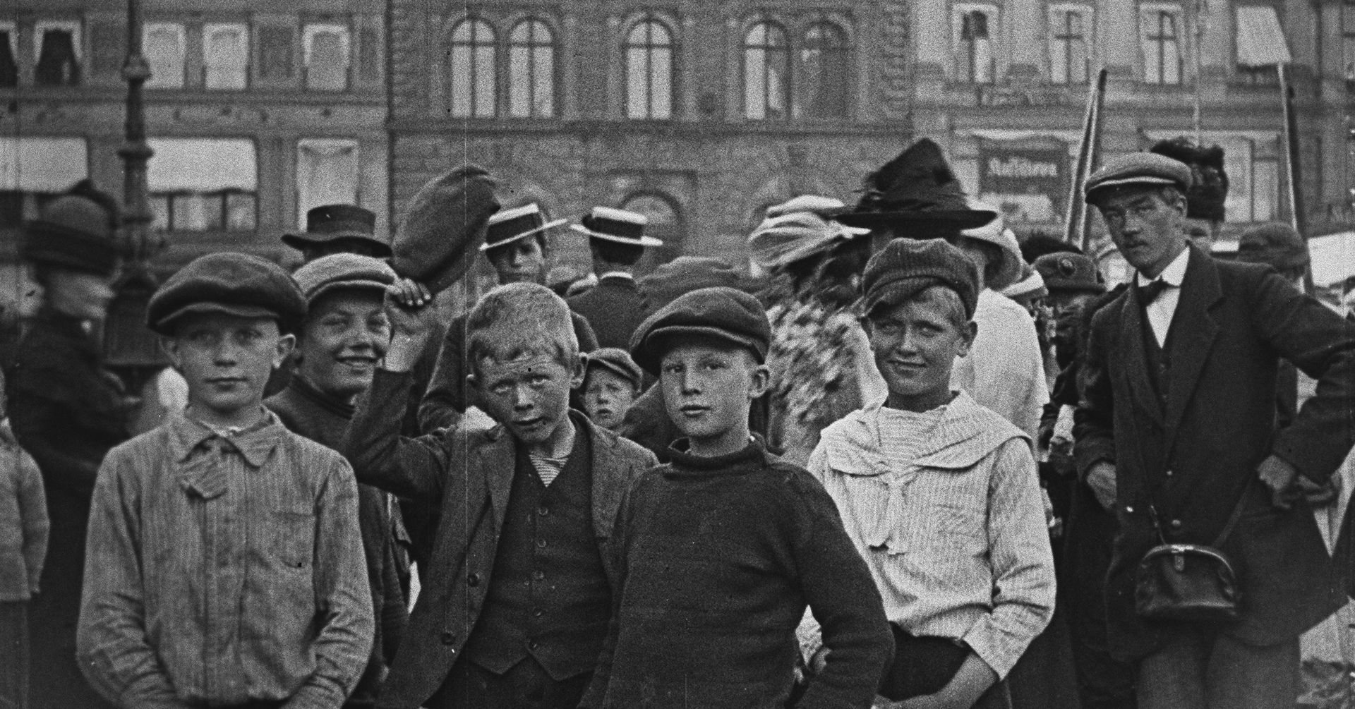 Et bilde som viser flere unge gutter i sixpense og arbeidsklær. De ser smilende og nysgjerrige ut på oss. De står foran en større murbygning, og på høyre side står det noen menn i dress og hatter og også sixpenser.