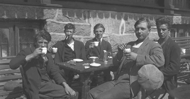 Fem unge menn sitter sammen ved en kafé ute, og nyter kaffe og sigarett. De myser mot sola.