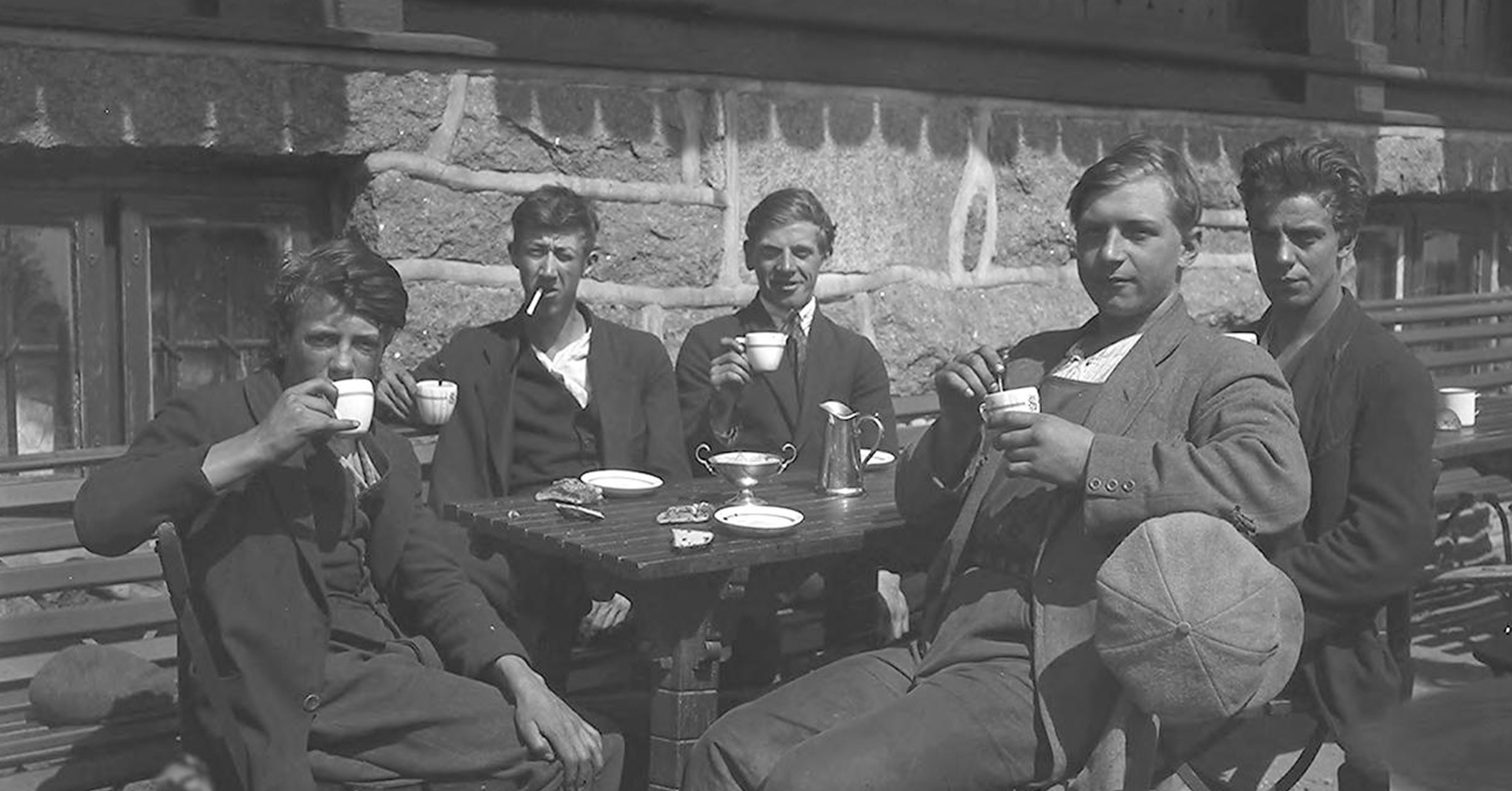 Fem unge menn sitter sammen ved en kafé ute, og nyter kaffe og sigarett. De myser mot sola.