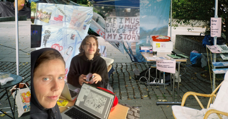 To unge kvinner ser mot oss. De sitter utendørs under et telt, omgitt av plakater og ser ut til å jobbe med utklipp til en avis