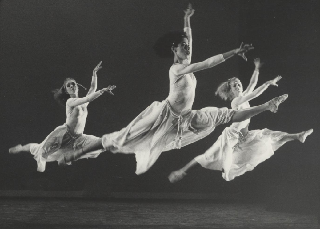 Tre hvitkledde ballettdansere hopper med bena utstrakt og hendene i været.