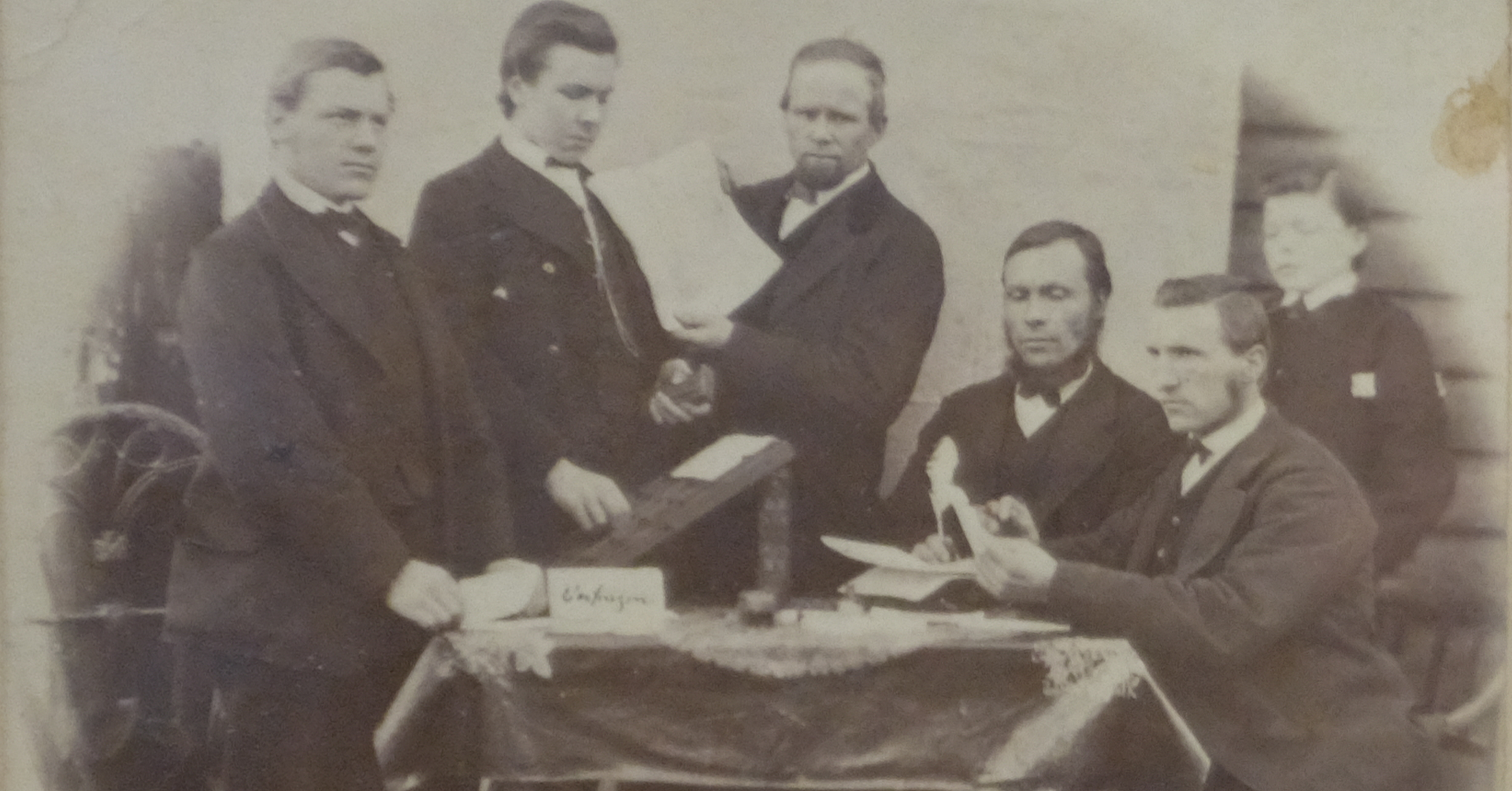 5-6 pent kledde menn står og sitter rundt et bord. Bildet er gammelt, og på bordet ligger det noe som ser ut som brev