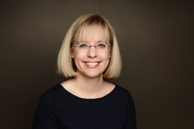 Kvinne med lyst hår og briller som smiler.