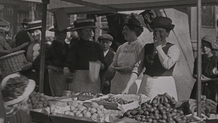 Flere kvinner i forkle og hatter har seg en god latter ved noe som ser ut som et matmarked.