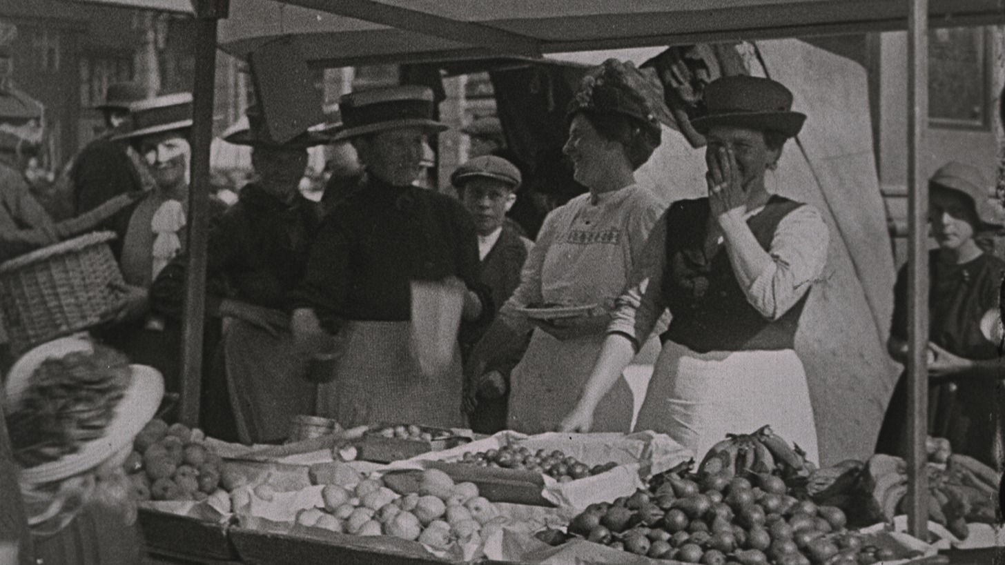 Flere kvinner i forkle og hatter har seg en god latter ved noe som ser ut som et matmarked.