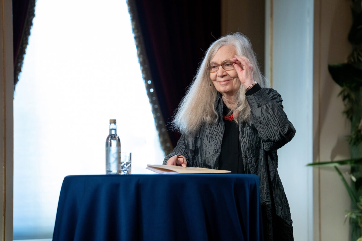 En blid dame med grått, langt hår står ved et podium og retter på brillene some. På bordet foran henne står en flaske vann og noen glass. I hånden holder hun noen ark.