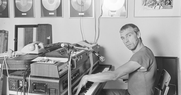 En ung mann sitter foran et avansert keyboard med mikrofoner og ledninger. På toppen av keyboardet er det sigarettsneiper og en telefon. På veggen ser man flere platinumplater i rammer. Den unge mannen spiller på keyboardet og ser nysgjerrig på oss.